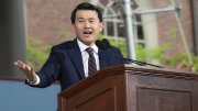 Ronny Chieng wearin a suit speaks passionately at a wooden podium outdoors.