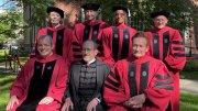 Group of seven individuals in academic regalia, smiling outside on a sunny day.