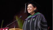 A smiling woman in academic regalia speaks at a podium during a graduation ceremony.