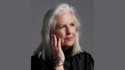 A woman with long, silver hair rests her chin on her hand, wearing a black top.