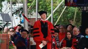 A speaker in academic regalia stands before an audience during a ceremony outdoors.