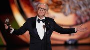 A man in a tuxedo gestures while holding an award on a stage.