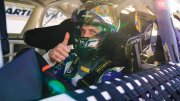 Racing driver gives a thumbs up from inside a car, wearing a helmet and safety gear.