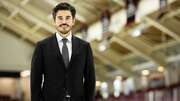 A man in a suit stands confidently on an ice rink, smiling, with a sports arena in the background.