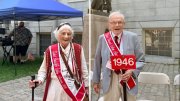 Older woman in a white outfit with a cane and red sash, alongside an elderly man in a gray suit holding a sign.