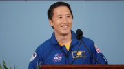 Smiling astronaut in blue jumpsuit speaking at a podium, colorful background.