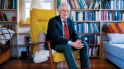 Harvey Mansfield seated in a bright yellow chair, surrounded by bookshelves and cozy decor.