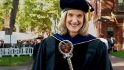 Katie O’Dair in academic regalia holds a ceremonial staff outdoors at a graduation ceremony.
