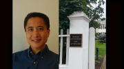 A man smiling in a blue shirt next to a white gate with a plaque and greenery.