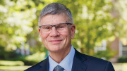A smiling William C. Kirby in a suit with glasses stands outdoors, surrounded by greenery.
