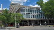 Modern building with large glass windows, surrounded by greenery and pedestrians.