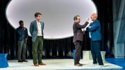 Three actors perform on stage, with one actor interacting closely with another, set against abstract backgrounds.