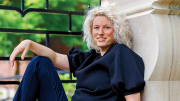 A woman with curly blonde hair sits casually against a stone wall, wearing a black top and jeans.