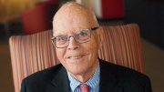 A smiling elderly man in glasses sits comfortably in a striped armchair.