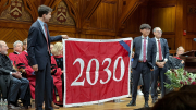 Two men hold up a red banner with "2030" written on it in white in a wood paneled room
