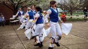 Morris dancers in white trousers and colorful vests wave handkerchiefs as they dance.