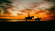 Photograph of two riders on horseback crossing a plain, silhouetted against the setting sun