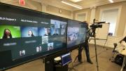 A hybrid classroom showing the large screens enabling a teacher to see all the students, remote or present, as he is followed by a camera operator.