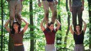 Four people perform a balancing act in a forest, with two standing on shoulders.