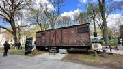 A red-brown train car with boarded-up windows, harsh metal siding, and faded industrial German lettering is on display in Harvard Yard.
