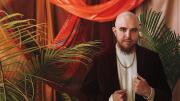 A formal portrait or José Olivarez in a gold chain and velvet suit jacket, standing amid red drapes and palm plants.