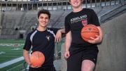 Two students pose in Harvard Stadium. One holds a basketball and the other holds a soccer ball.
