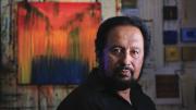 Painter George Oommen in his studio. His painting <em>Visions of Kerala 2</em> (acrylic on  canvas, 2005) hangs behind him. 