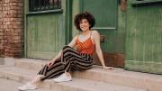 Adriana Colón, a casually dressed, smiling young woman with curly brown hair, sits in a doorway.