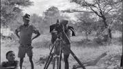 “/Gaishay focusing the camera,” a photograph taken during a 1957-58 ethnographic expedition in Namibia, shows three indigenous boys, one standing behind a camera on a tripod