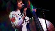 esperanza spalding, wearing a white jumpsuit with a "Life Force" logo, stands next to an upright bass