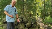 A white haired professor emeritus standing by a stone wall in the woods explaining its origins 