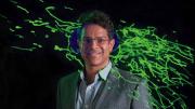 Vamsi Mootha with an image from his lab showing thread-like mitochondria (green) moving within a cell.