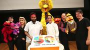 Two bakers present a large birthday cake, surrounded by Muppets and their puppeteers.