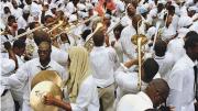 A street crowd of black men and women, all dressed in white, either playing or responding to the playing of dozens of trombones