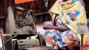 A grinning woman in traditional Nigerian dress sits cross-legged on the floor surrounded by modern devices, including a power strip, a land-line telephone, and a desktop computer displaying on its screen a duplicate image of the entire montage.