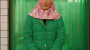 Photograph of a woman in a bright red-and-white hijab and bright green winter coat against a green curtain background, looking directly at the camera