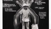 A man in frayed clothing stands in front of a mural marking a timeline of violent acts against immigrants. The mural's dates surround a large pair of hands, handcuffed together with rope.