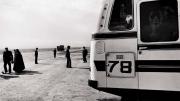 Black-and-white photo of a bus, dominating the right half of the photograph, while a handful of people, and a truck driving toward the horizon, appear as small figures in the left half.
