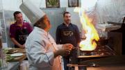 Two men look on while a chef cooks a flaming dish over a stove.