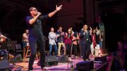 Singer Ali Sethi gestures toward the audience at Sanders Theatre, while students dance on stage behind him.