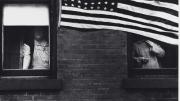 Robert Frank photograph of partially obscured people looking out of windows behind a large American flag