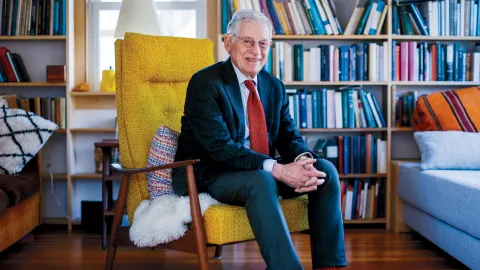 Harvey Mansfield seated in a bright yellow chair, surrounded by bookshelves and cozy decor.