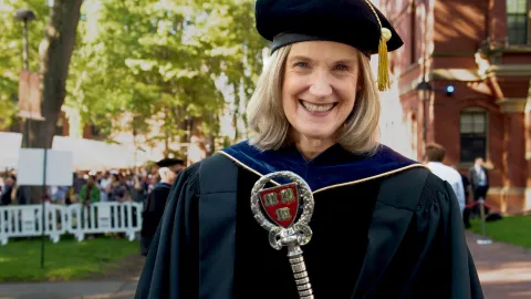 Katie O’Dair in academic regalia holds a ceremonial staff outdoors at a graduation ceremony.