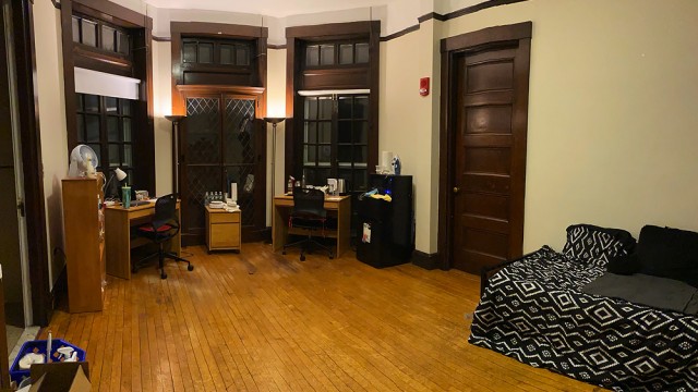 A large dorm room with desk, bed, lamps, and quarantine necessities, including bottled water, masks, and sanitizing supplies.