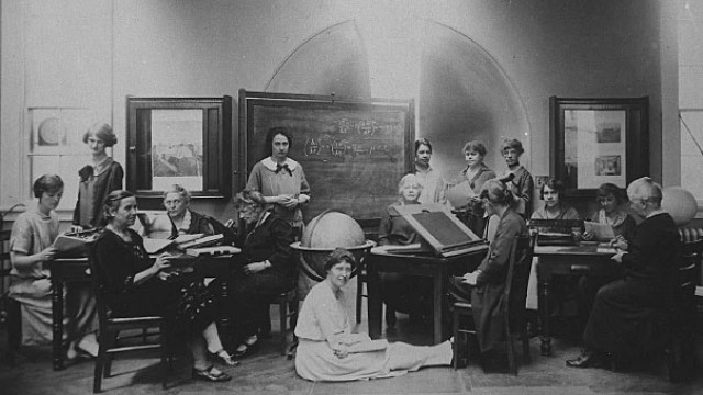 Several Harvard Computers and Radcliffe students pose for a photograph together, seated at desks and sitting on the floor