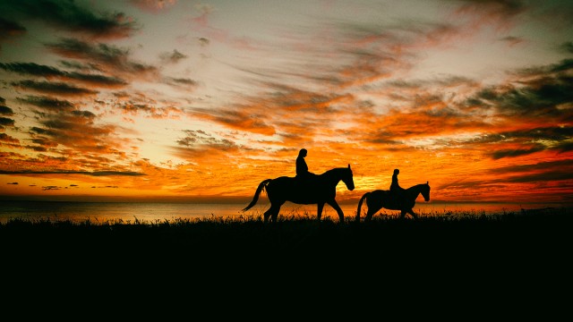 Photograph of two riders on horseback crossing a plain, silhouetted against the setting sun