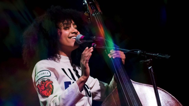 esperanza spalding, wearing a white jumpsuit with a "Life Force" logo, stands next to an upright bass