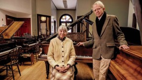An older couple standing in a large room filled with pianos