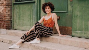 Adriana Colón, a casually dressed, smiling young woman with curly brown hair, sits in a doorway.