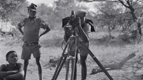 “/Gaishay focusing the camera,” a photograph taken during a 1957-58 ethnographic expedition in Namibia, shows three indigenous boys, one standing behind a camera on a tripod